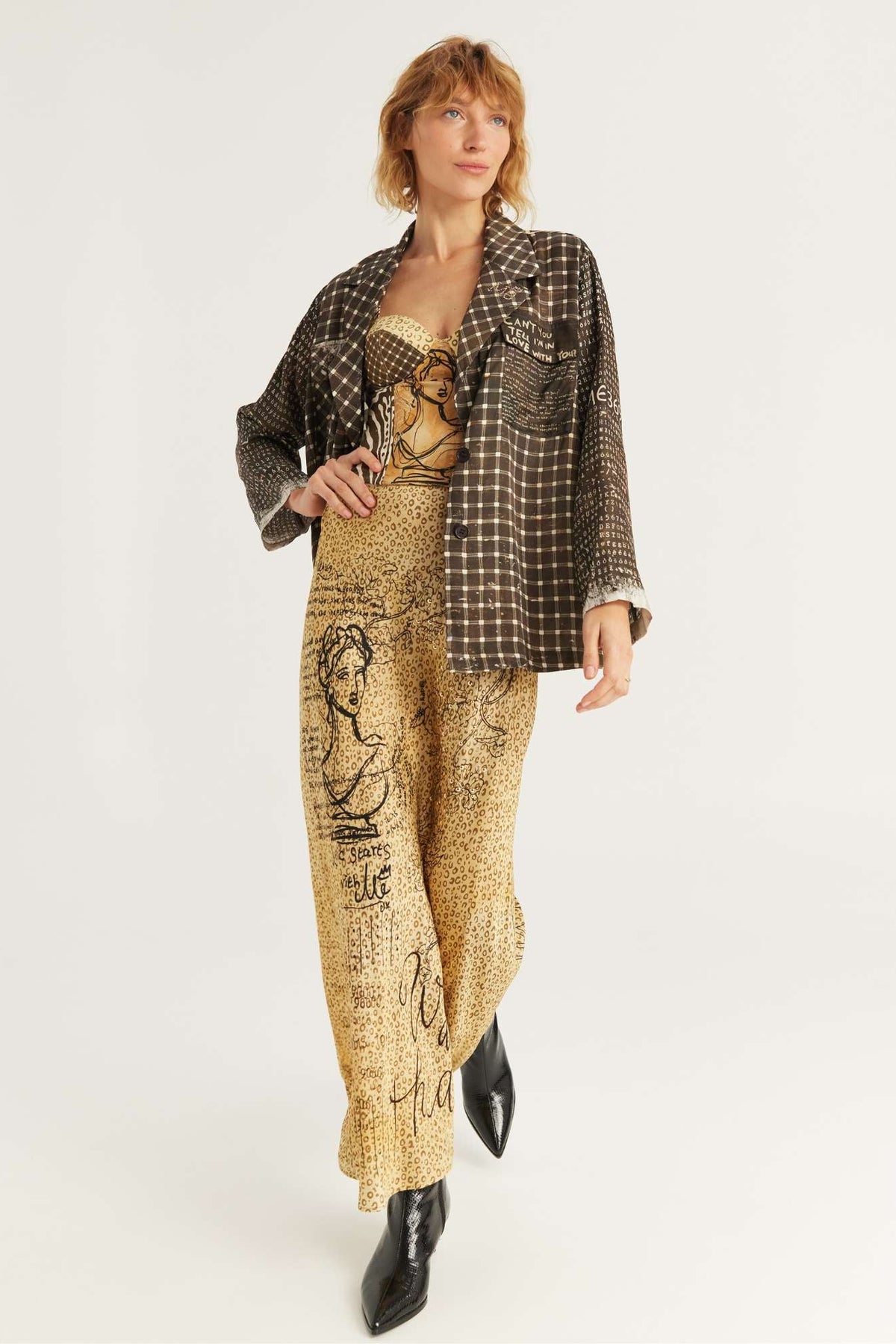 A model wearing a brown and black checkered plaid Audrey Collar Jacket in Noir Muse print over a beige and black graphic print jumpsuit, paired with black ankle boots, against a plain white studio background.