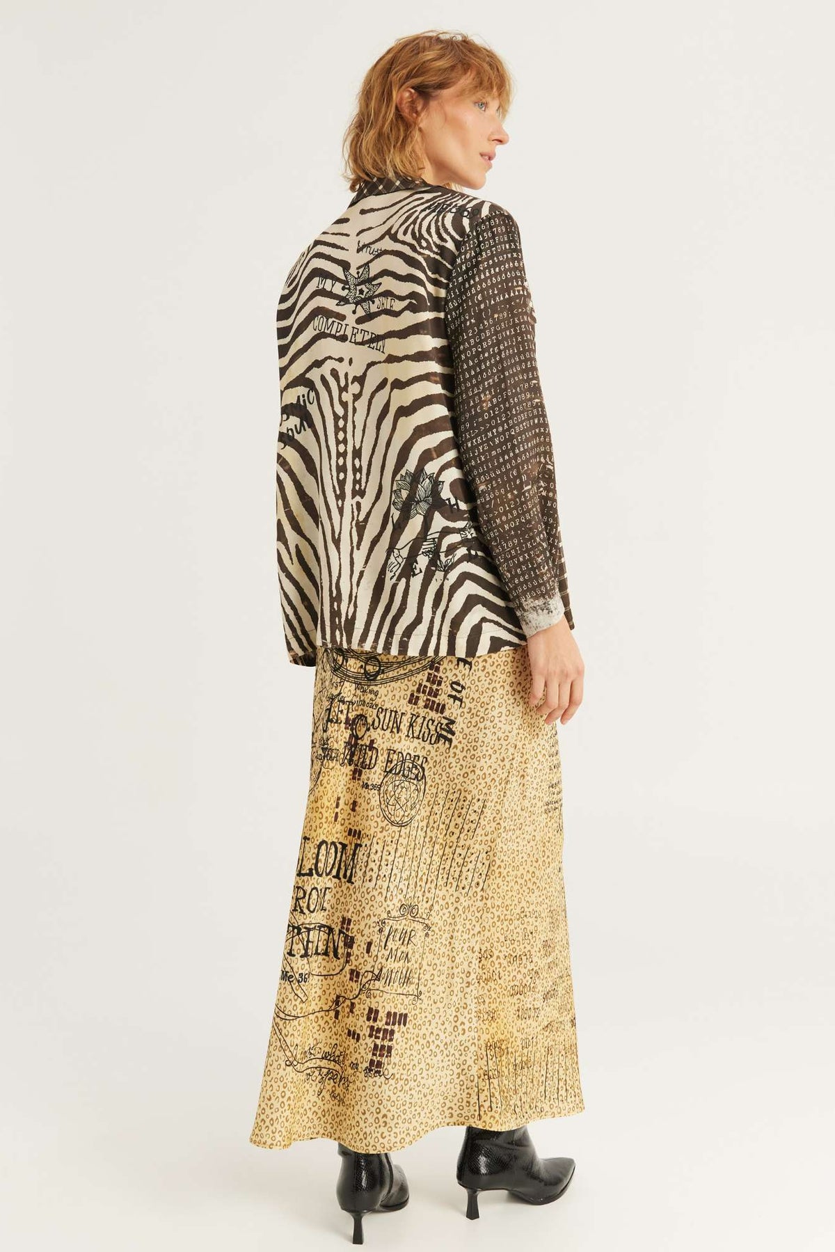A model shown from the back wearing a long-sleeved Audrey Collar Jacket with a split Noir Muse design: a black and beige zebra print on one side and a black mesh pattern on the other, paired with a beige skirt featuring black text and graphic prints, set against a plain light gray background.