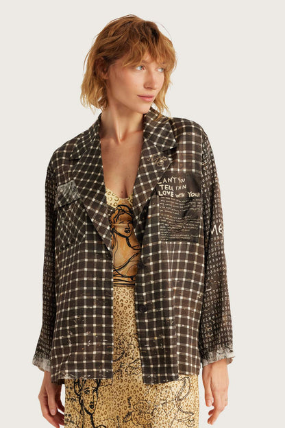 A model wearing a brown and black plaid open-front Audrey collar jacket in Noir Muse print with graphic text patches over a patterned top and skirt, set against a plain light gray background.