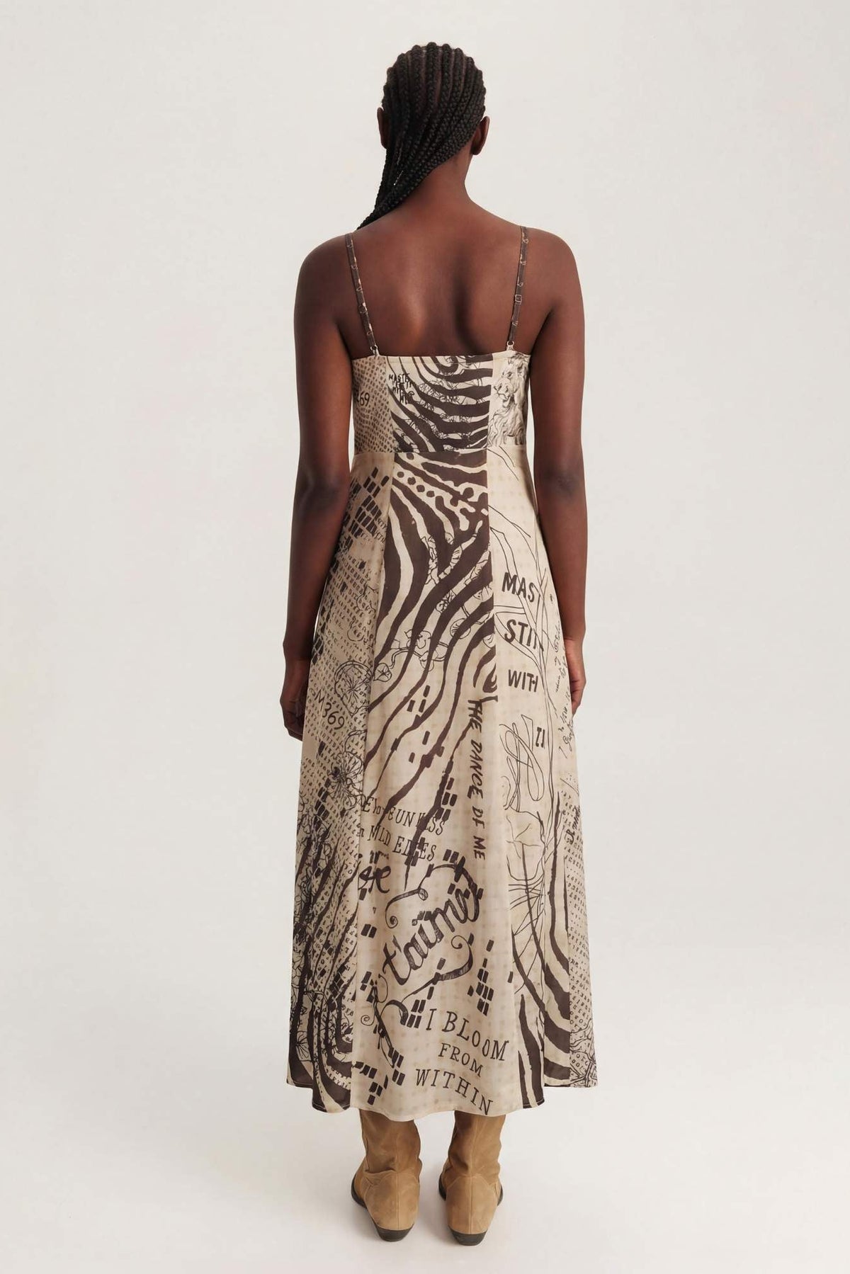 A woman viewed from behind wearing a sleeveless corset maxi dress with thin straps and an abstract black and beige print, paired with tan ankle boots, against a plain light background.