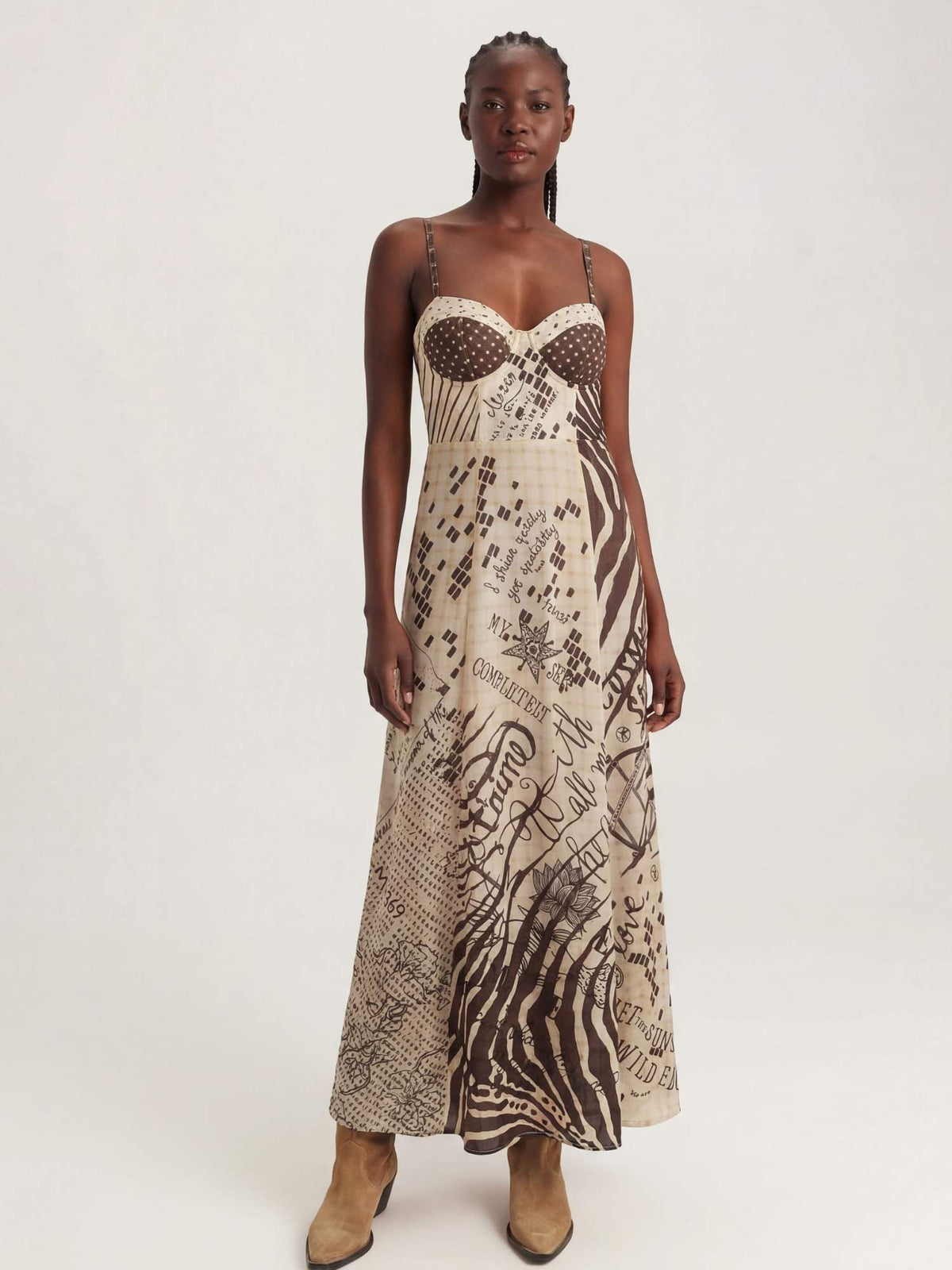 A model wearing a sleeveless corset maxi dress with a beige and brown abstract Noir muse print, featuring a sheer polka-dot bustier bodice and thin spaghetti straps, paired with tan ankle boots, against a plain light gray background.
