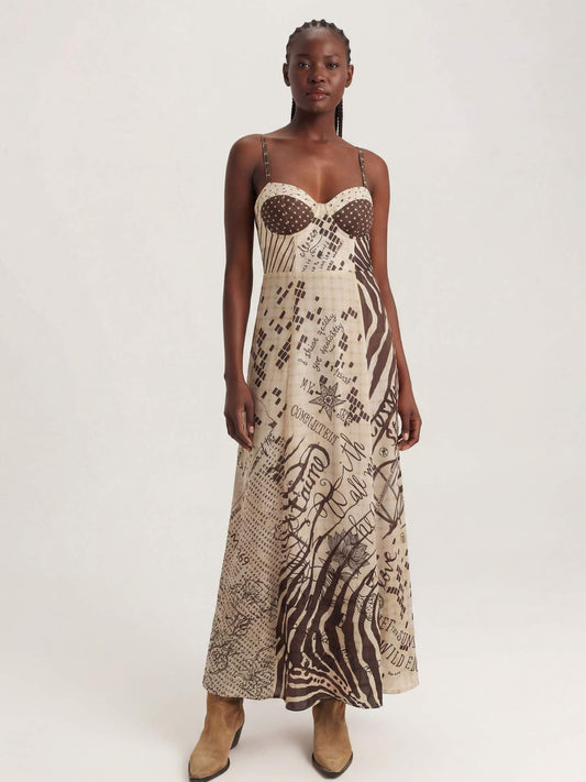 A model wearing a sleeveless corset maxi dress with a beige and brown abstract Noir muse print, featuring a sheer polka-dot bustier bodice and thin spaghetti straps, paired with tan ankle boots, against a plain light gray background.