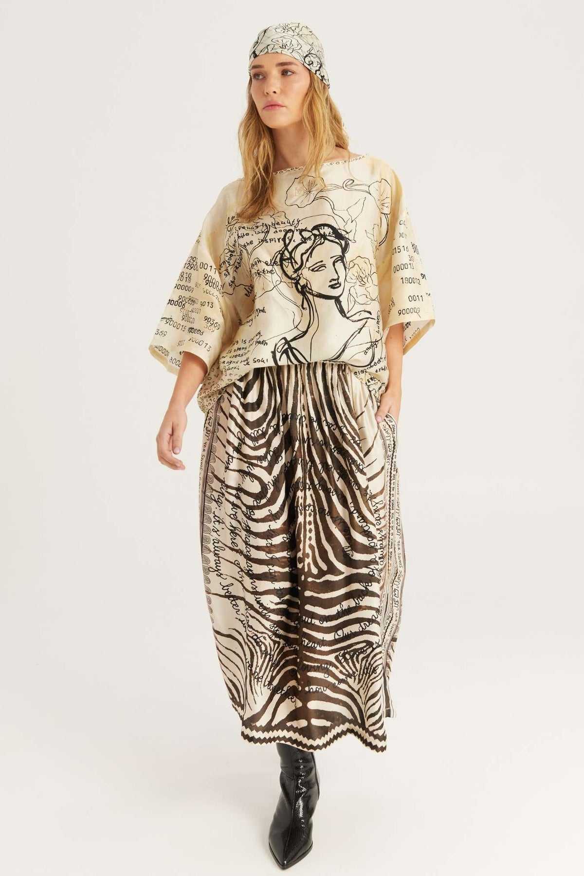 A model wearing a two-piece printed outfit consisting of a beige tunic top with black graphic illustrations and wide-leg skirt featuring a brown and black zebra Noir Muse print, paired with a matching patterned headscarf and black boots.