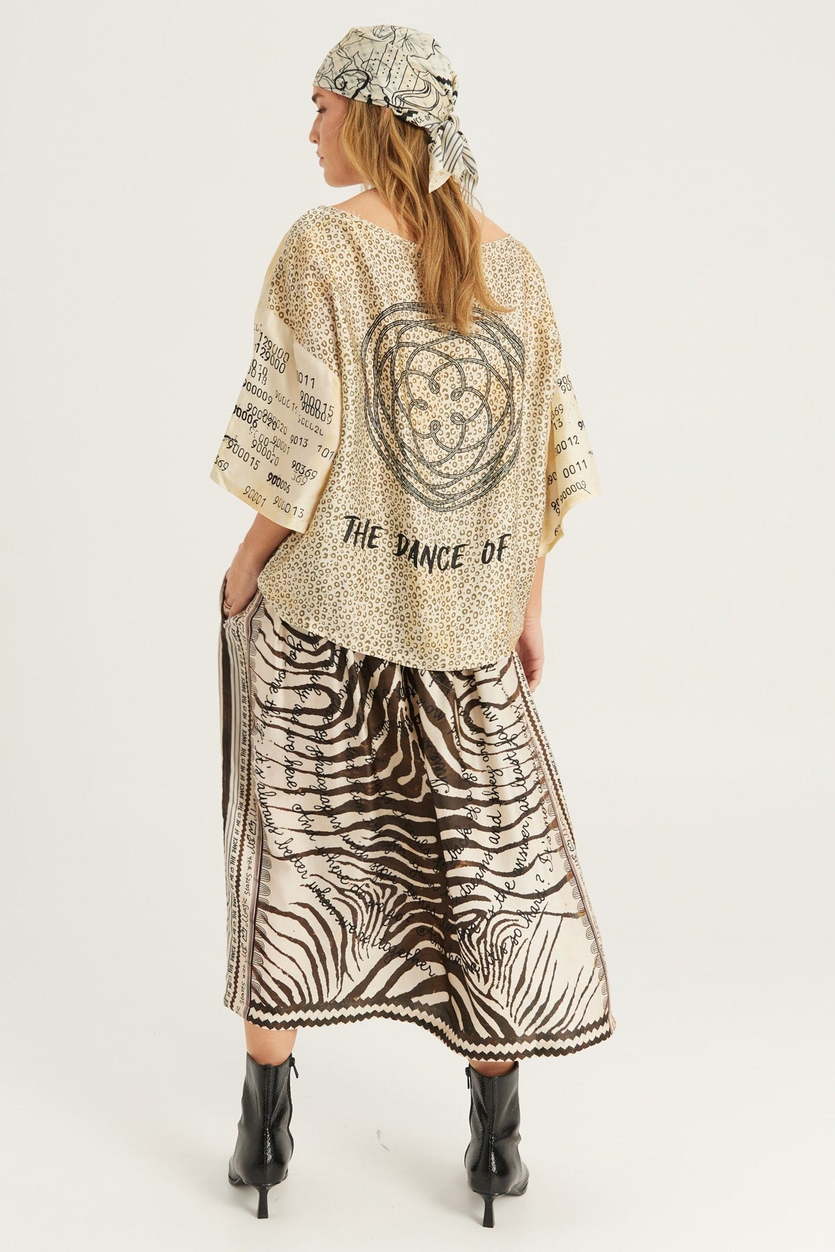 A model shown from the back wearing a beige and black patterned kaftan top with 'THE DANCE OF' text and a large circular graphic, paired with a matching zebra-print Noir Muse skirt and a patterned headscarf, on a light gray background.