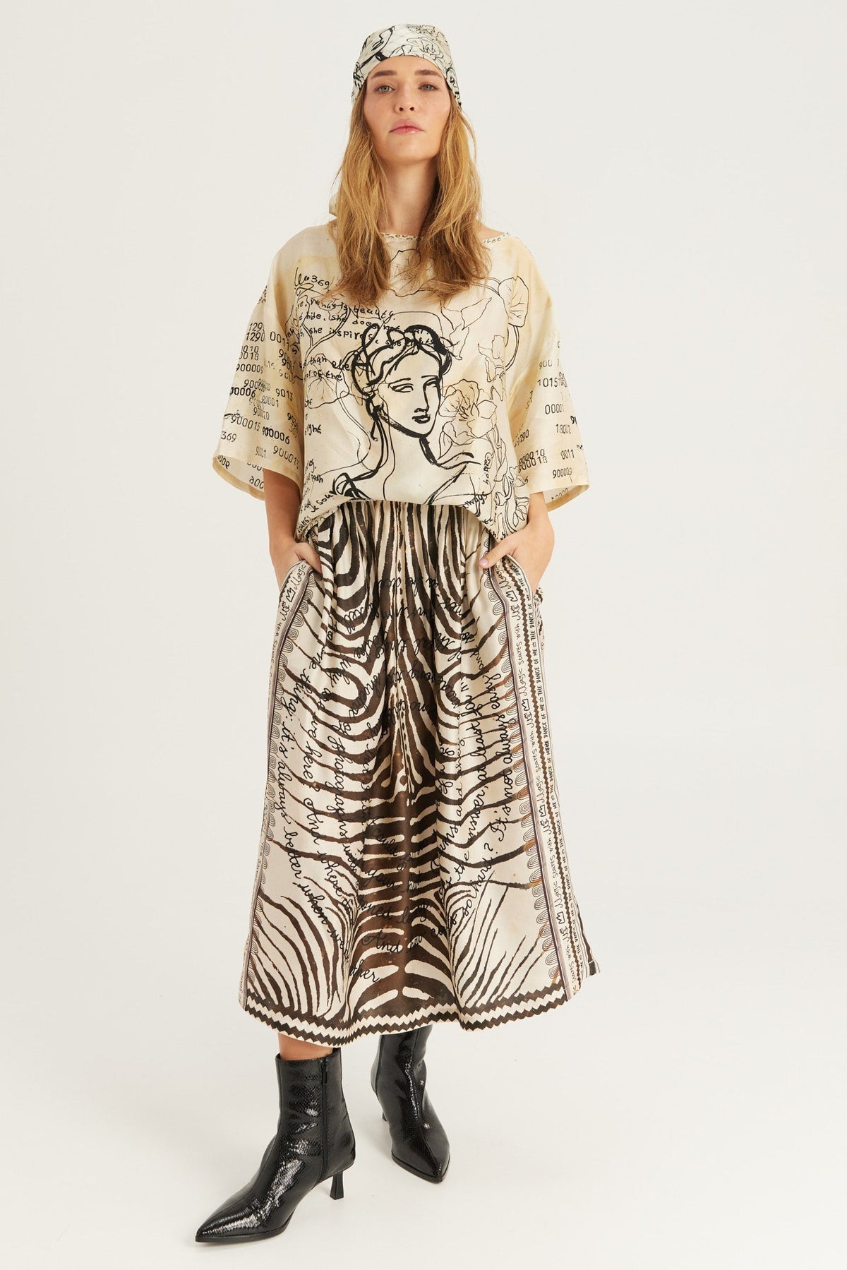 A model stands against a light gray studio background wearing a beige and black Noir Muse patterned two-piece outfit with a wide-sleeve top, matching skirt, black ankle boots, and a matching headscarf.