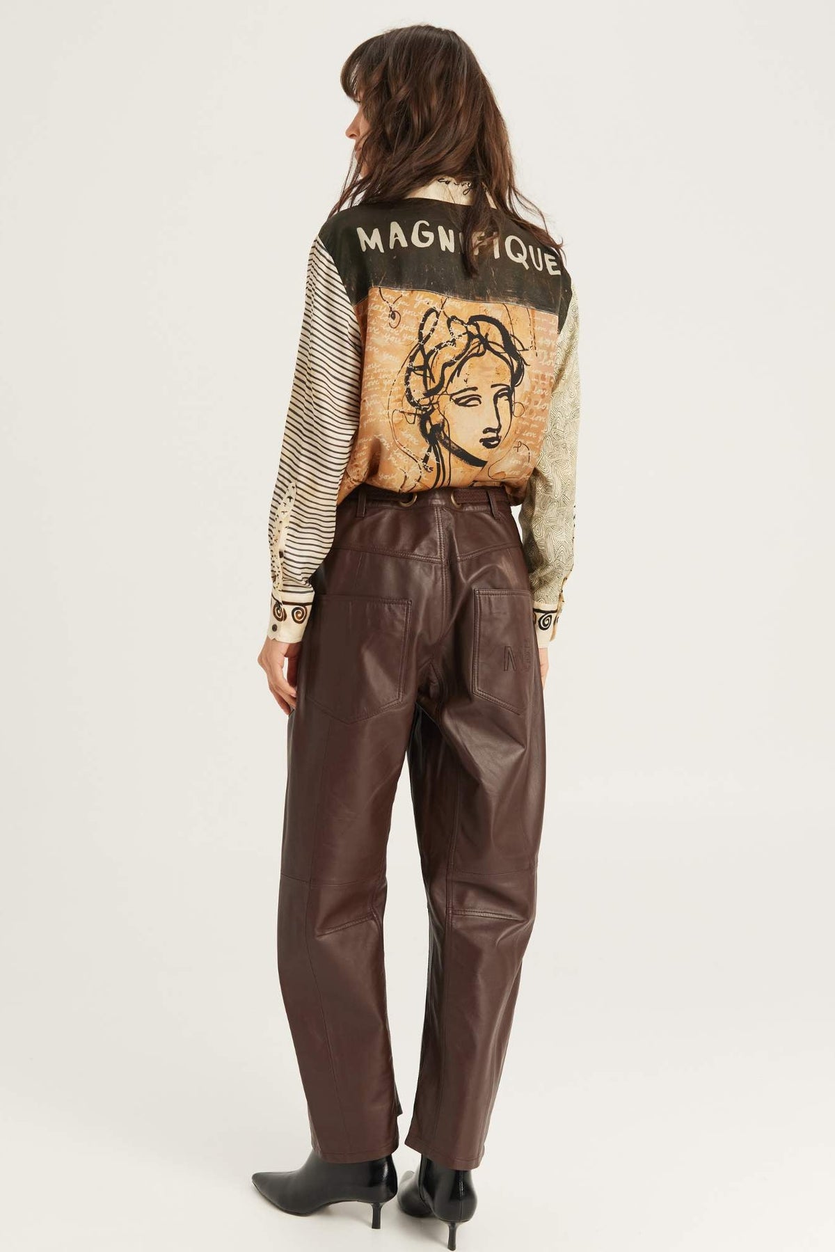 A model is shown from the back wearing a silk shirt with a vintage Noir muse art print on the back and striped sleeves, paired with dark brown faux-leather trousers and black ankle boots, against a plain white studio background.