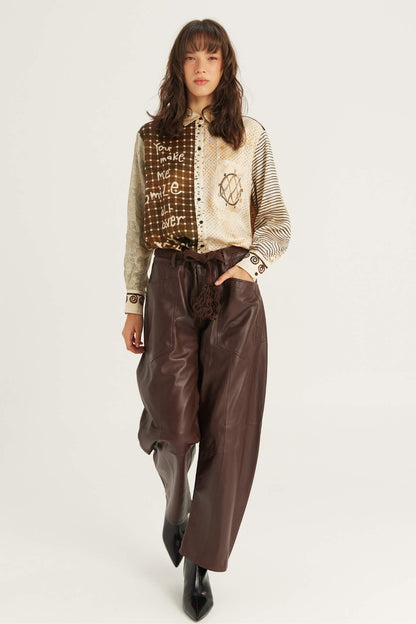 A model is shown from the head to the boots, wearing a beige and brown Noir Muse patterned button-up shirt with striped sleeves, paired with dark brown wide-leg faux-leather pants and black pointed-toe boots, against a plain white studio background.