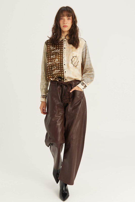 A model is shown from the head to the boots, wearing a beige and brown Noir Muse patterned button-up shirt with striped sleeves, paired with dark brown wide-leg faux-leather pants and black pointed-toe boots, against a plain white studio background.