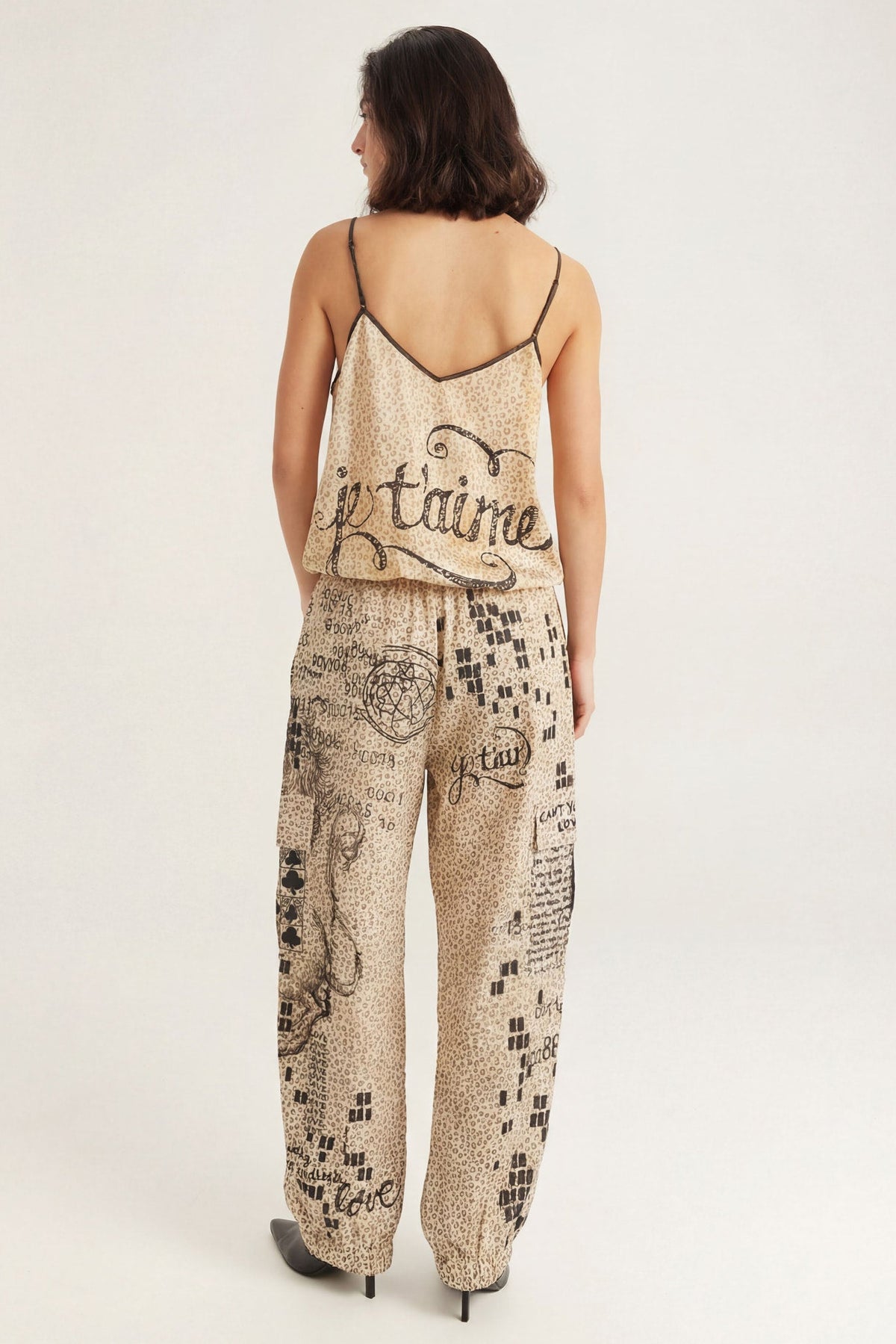 A woman shown from the back wearing a beige and black patterned camisole top with 'je taime' written across the back and matching cargo pants with a black graphic Noir Muse print, set against a plain light gray studio background.