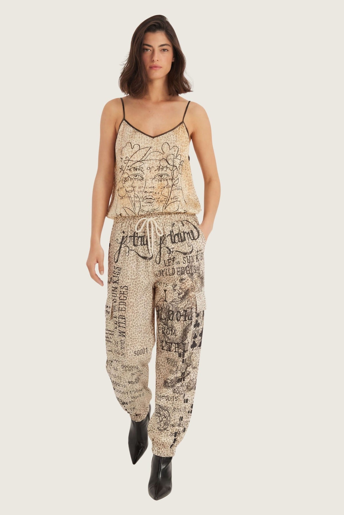 A woman modeling a matching beige and black graphic Noir Muse  print camisole top and cargo pants set, paired with black pointed-toe ankle boots, against a solid light gray background.