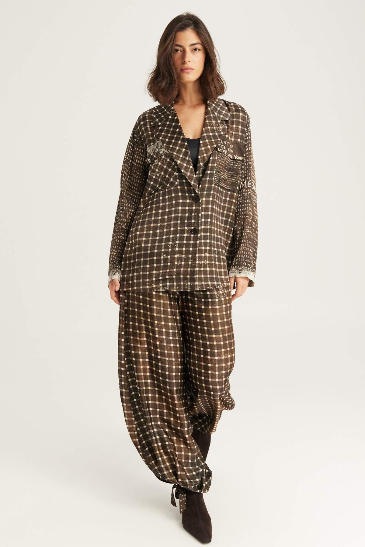 A model wearing a brown and beige checkered Noir Muse two-piece set consisting of a button-up shirt and matching Isla balloon pants, paired with dark brown ankle boots, against a plain light gray background.