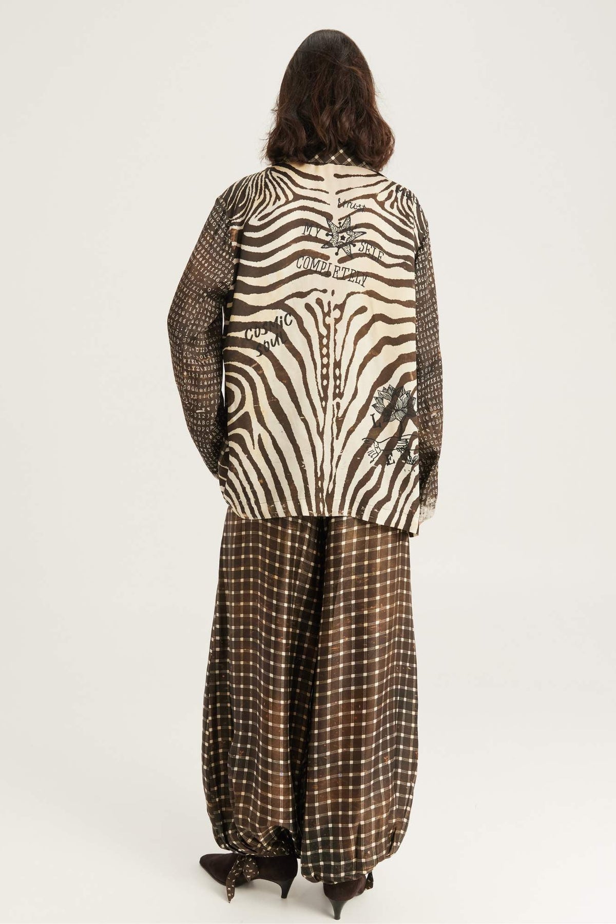 A model viewed from the back wearing a zebra/noir muse print jacket with text details and Isla balloon pants in noir muse print set against a plain white studio background.