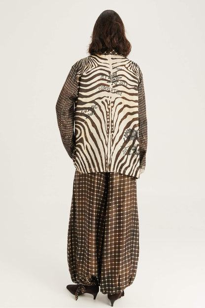 A model viewed from the back wearing a zebra/noir muse print jacket with text details and Isla balloon pants in noir muse print set against a plain white studio background.