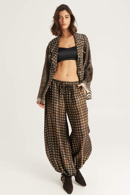 A model stands against a light gray studio background wearing a matching brown and beige checkered blazer and Isla balloon pants in Noir Muse print, styled with a black bandeau top and dark brown ankle boots.