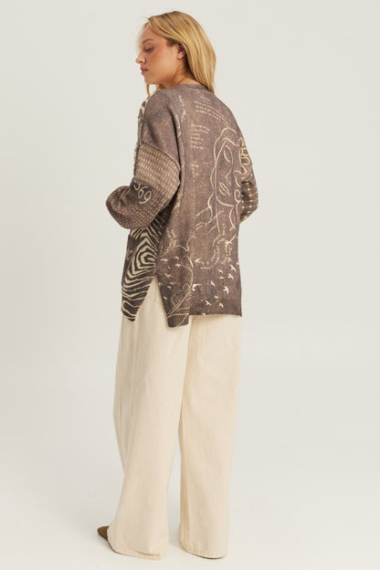 A woman shown from the back wearing a brown Noir Muse patterned Liv Long cardigan with abstract line drawings and textured knit sleeves, paired with cream wide-leg trousers, against a plain light gray studio background.
