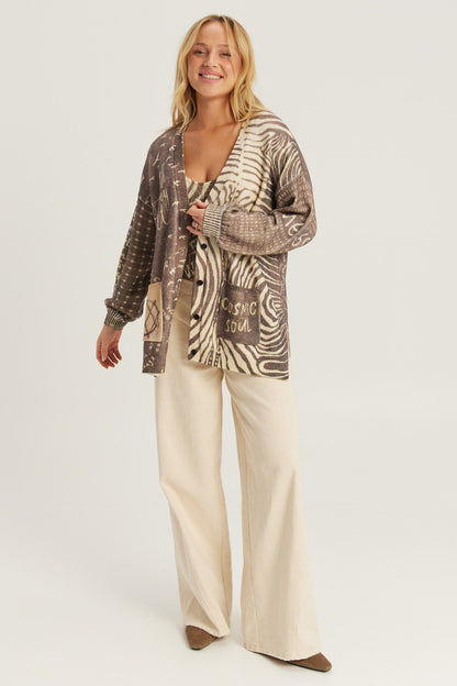 A woman modeling a mixed-media Noir Muse cardigan with abstract and zebra-like patterns in brown, beige, and black, paired with cream wide-leg trousers against a light gray studio background.
