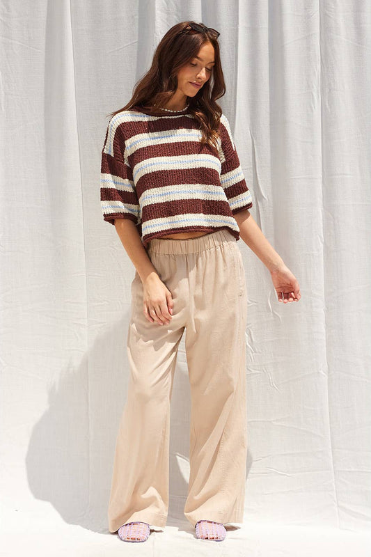 A woman modeling a brown and ecru striped cropped knit sweater with beige wide-leg pants, standing against a white draped fabric backdrop.
