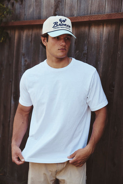 Man wearing a white t-shirt and cap with a Bronco brand logo against a wooden fence.