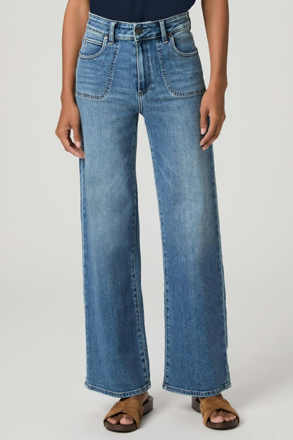 A model wearing a pair of high-waisted, Anessa Rounded Stitch wide-leg blue denim jeans with a monrovia faded wash, paired with brown slide sandals, shown against a light gray studio background.