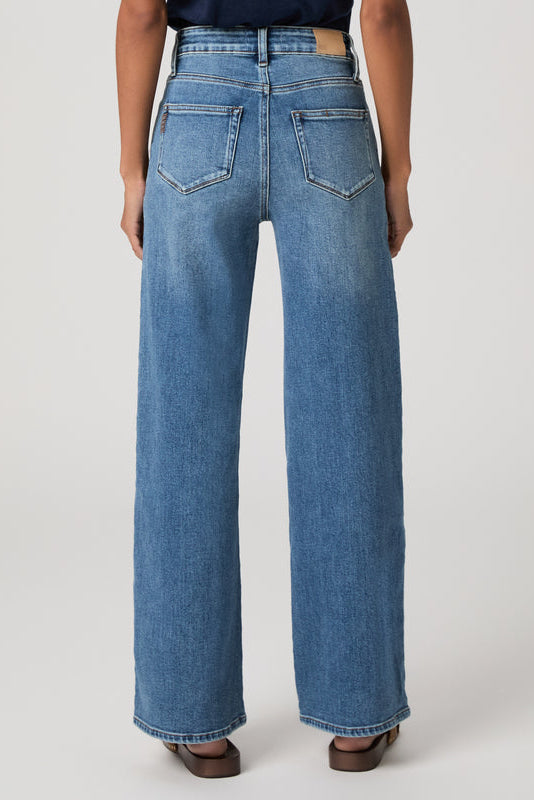 A person shown from the back wearing light-to-medium wash blue wide-leg Anessa Rounded Stitch jeans in Monrovia color variant, paired with a dark top and brown flat shoes, against a plain light gray studio background.