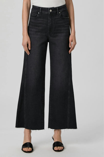 A model wearing a pair of wide-leg Anessa cropped jeans in ink black denim, paired with black slide sandals, shown against a neutral light gray studio background.