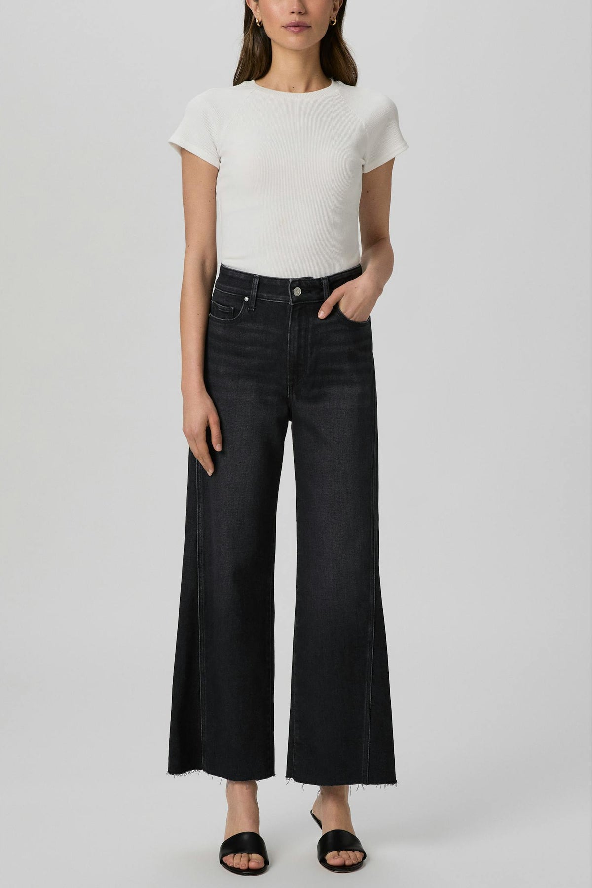 A model wearing a white short-sleeve crewneck top and Anessa ink black wide-leg cropped jeans with side slits, paired with black slide sandals, against a light gray studio background.