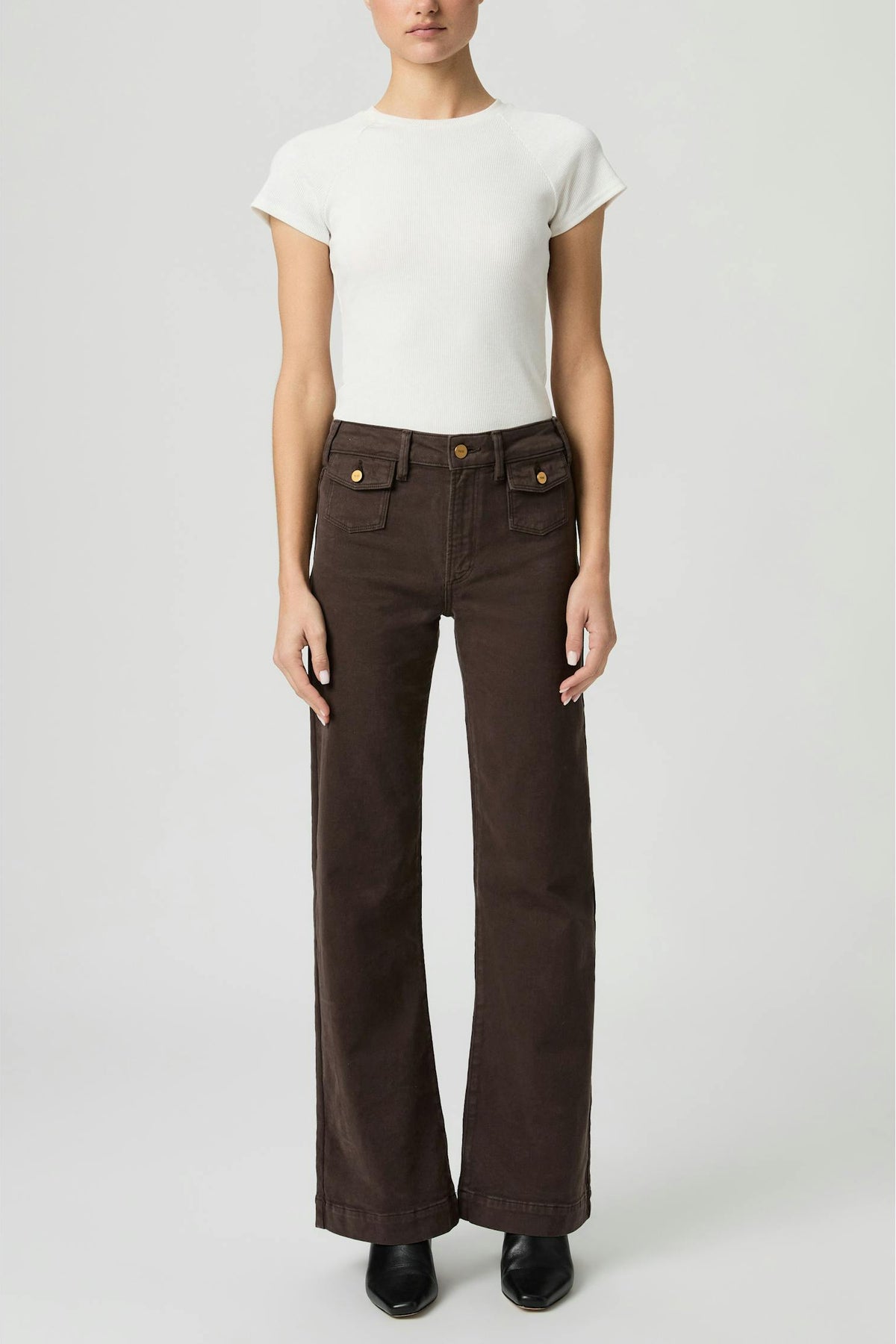A model wearing Leenah rich cholcolate brown wide-leg jeans with gold-tone button and pocket details, paired with a white short-sleeve top and black shoes, against a light gray studio background.