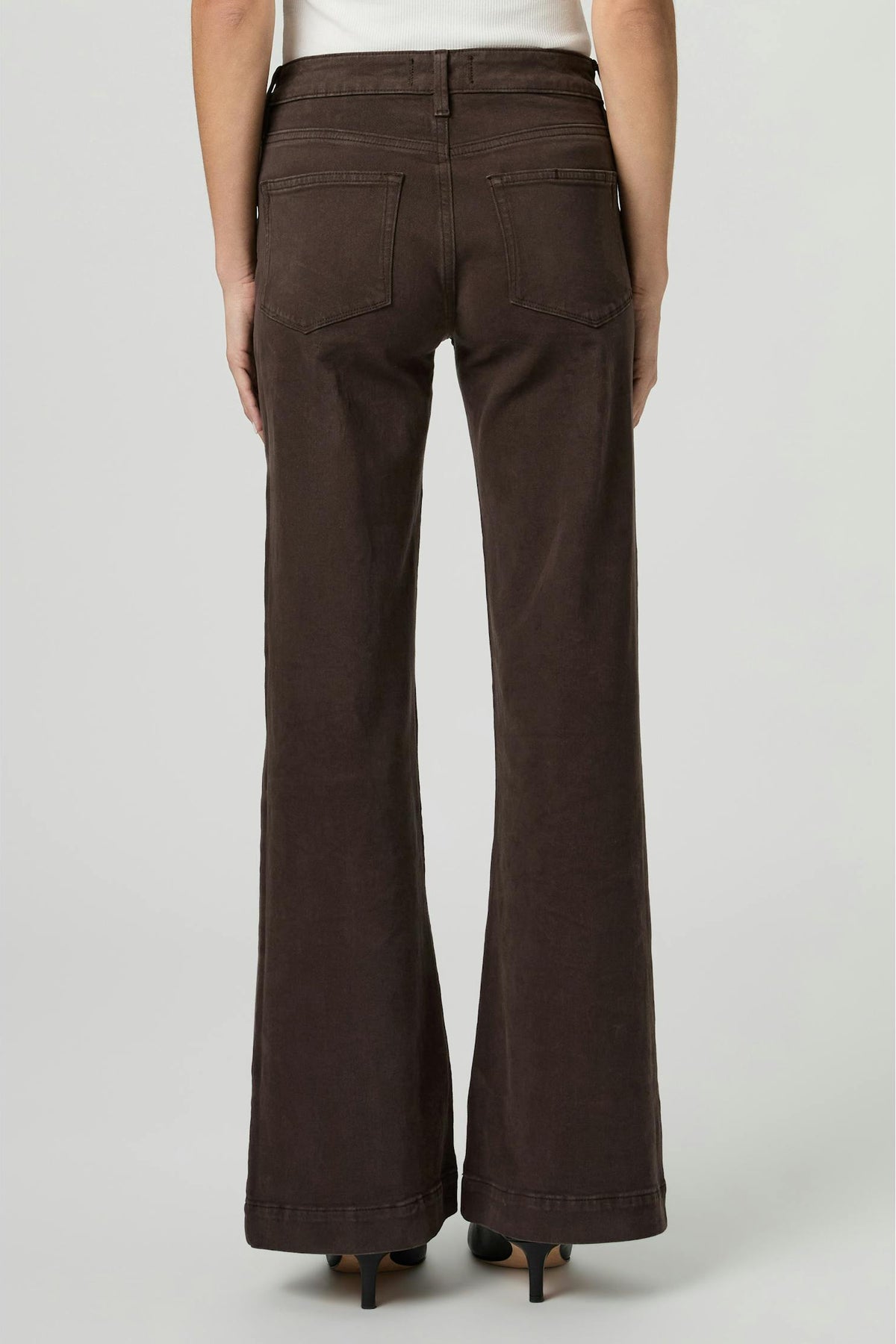 A person shown from the back wearing Leenah rich chocolate brown wide-leg flare jeans, paired with a white top and black heels, against a light gray studio background.