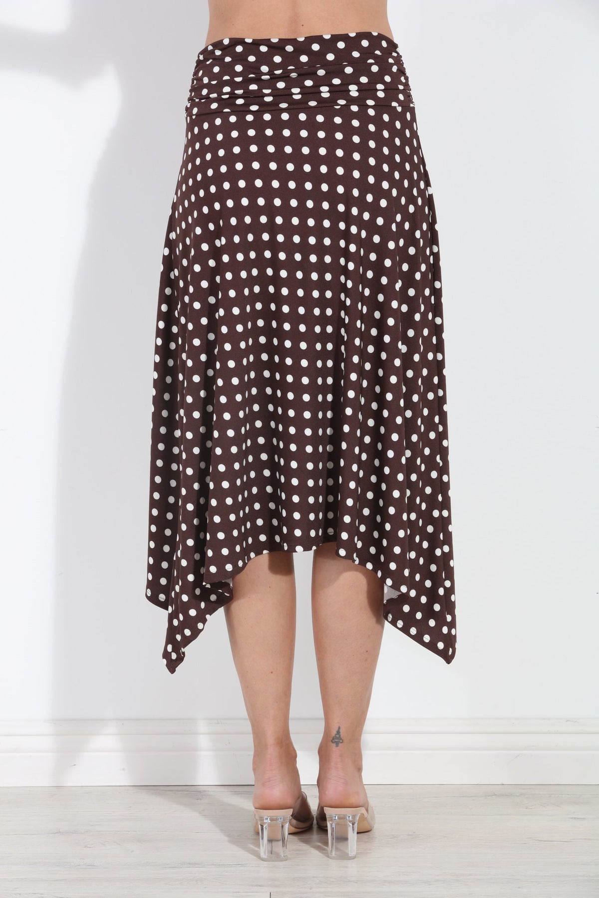 A brown and white polka dot high-low midi skirt worn by a model, shown from the back against a white wall.