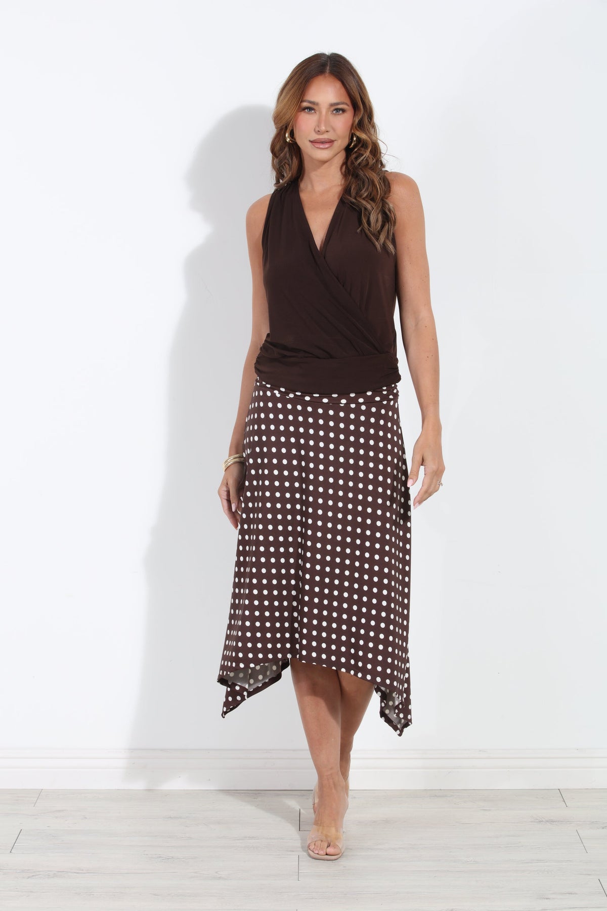 A woman modeling a sleeveless dark brown wrap-style top and a matching dark brown charlie bias cut midi skirt with white polka dots, standing against a plain white wall.