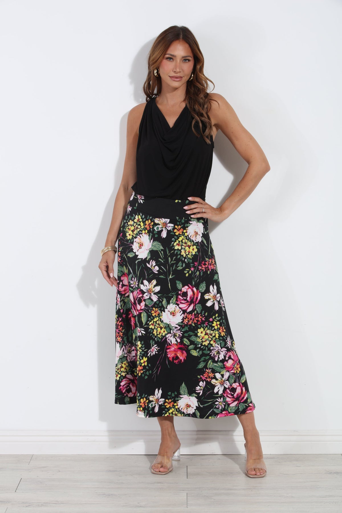 A woman modeling a sleeveless black cowl-neck top paired with a black midi skirt featuring a vibrant multicolor floral print, standing against a white wall.