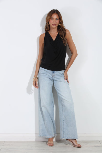A woman modeling a black sleeveless draped top and light-wash wide-leg jeans against a plain white wall.