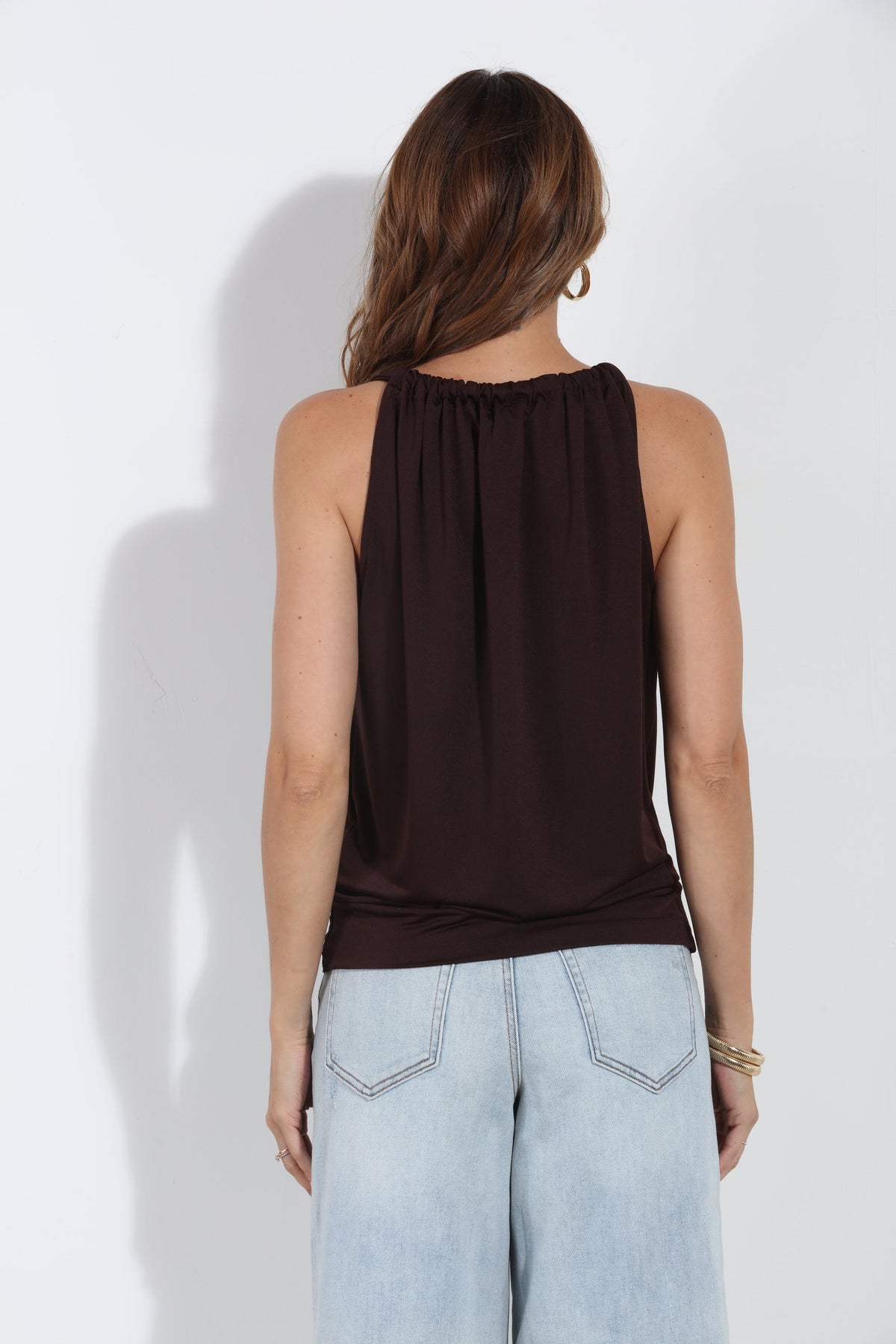 A woman shown from the back wearing a sleeveless dark brown top and light blue jeans, standing against a plain white wall.