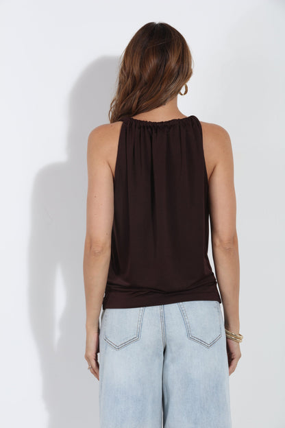 A woman shown from the back wearing a sleeveless dark brown top and light blue jeans, standing against a plain white wall.