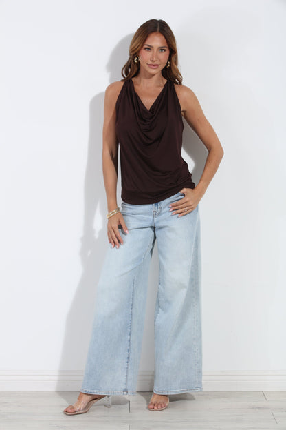 A woman modeling a dark brown cowl-neck sleeveless top and light-wash wide-leg jeans against a plain white wall.