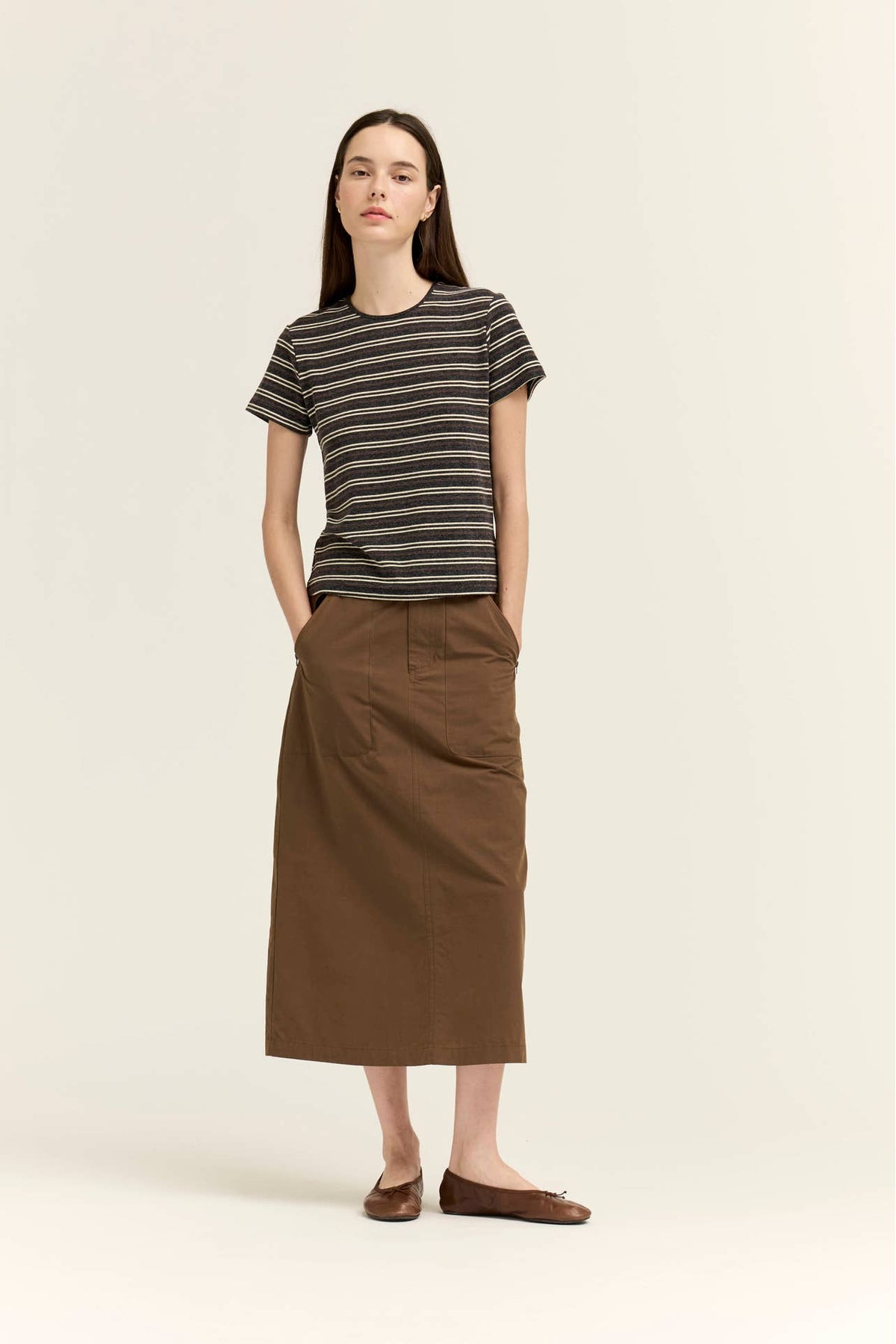 A woman stands against a plain light beige studio background wearing a short-sleeve crew-neck t-shirt with thin dark charcoal horizontal stripes and a long brown midi skirt.
