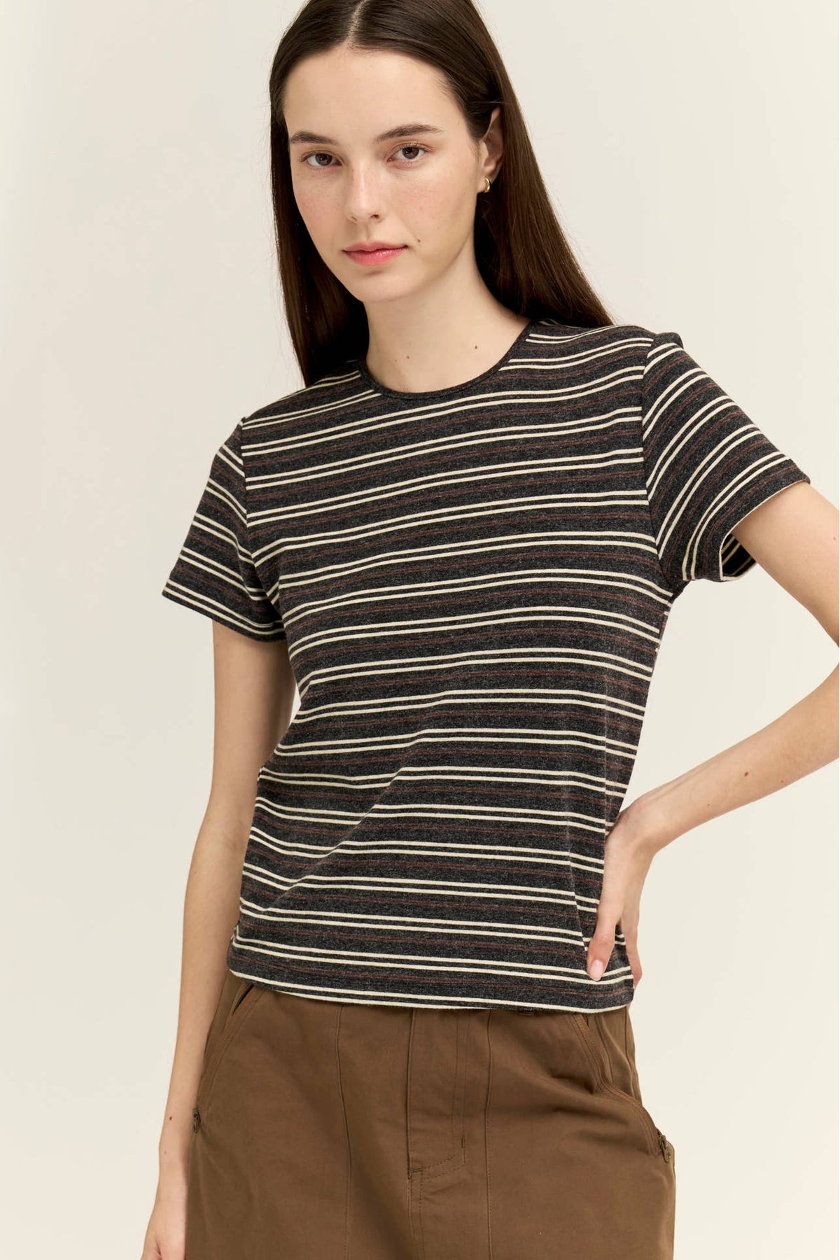A woman wearing a short-sleeve crewneck t-shirt with thin horizontal charcoal stripes, paired with brown pants, against a plain light beige background.