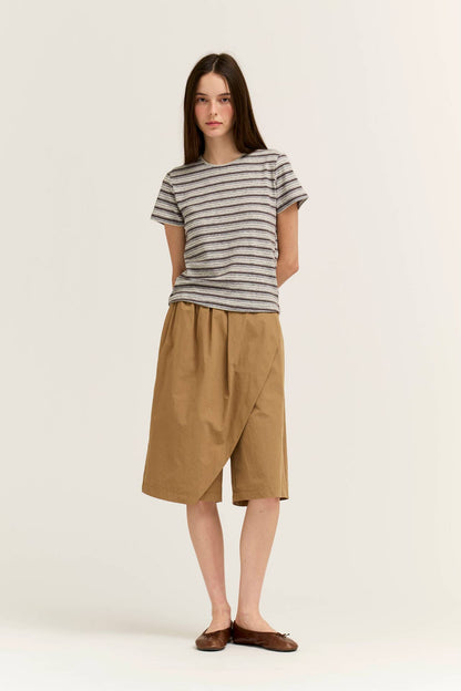 A woman modeling a short-sleeve light grey and white striped t-shirt paired with tan wrap-style knee shorts and brown flat shoes, standing against a plain light beige studio background.