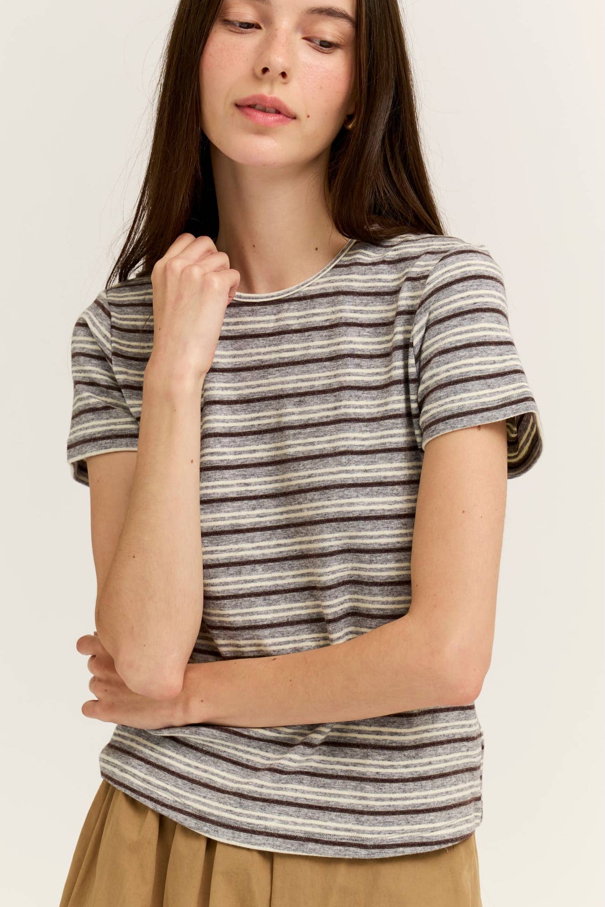A woman wearing a short-sleeve crewneck t-shirt with thin horizontal light grey and beige stripes, paired with a tan skirt, set against a plain light beige background.