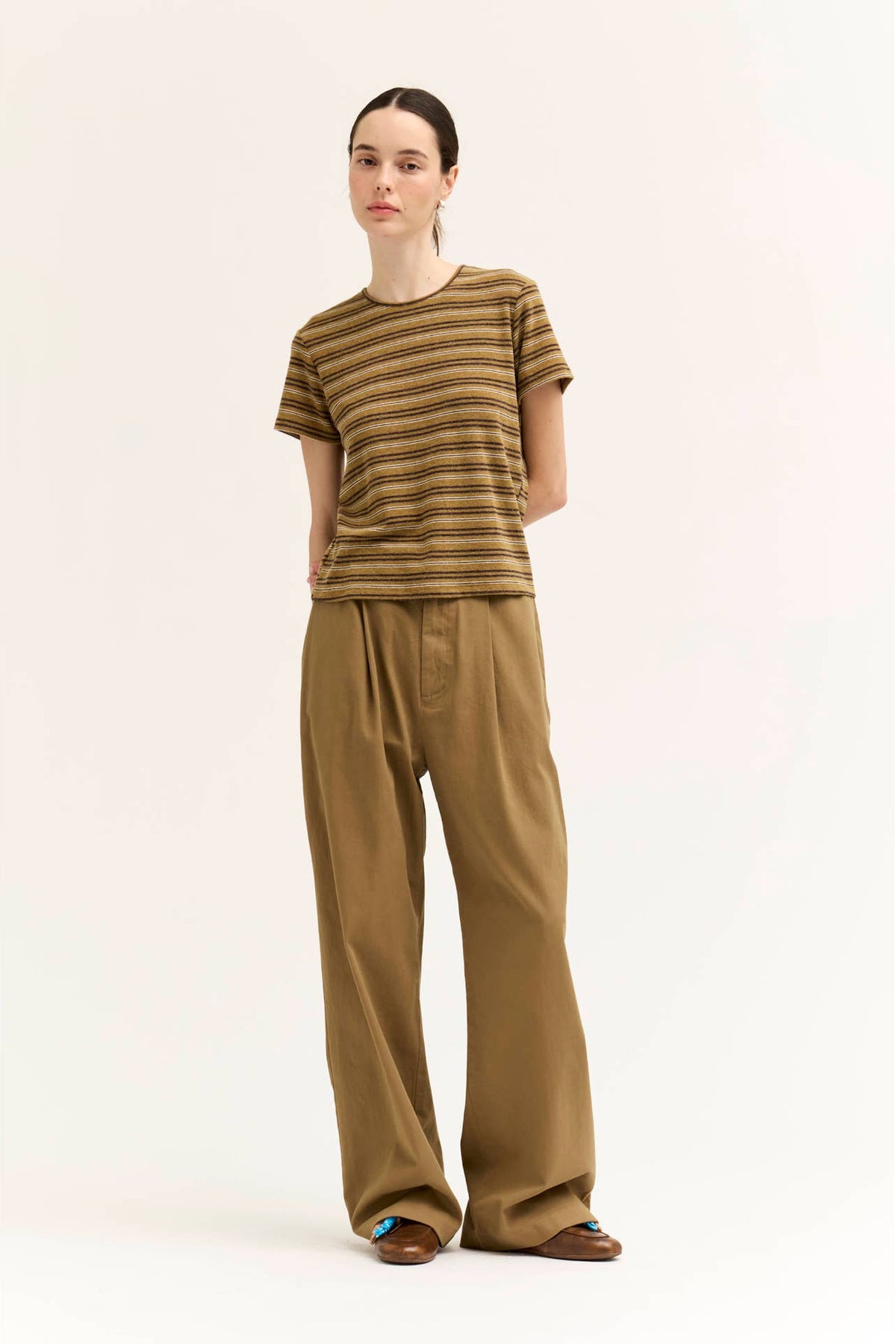 A model stands against a plain off-white studio background wearing a short-sleeve crew-neck t-shirt with horizontal "spice" color variant stripes, paired with wide-leg brown trousers and brown sandals.