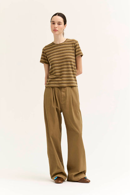 A model stands against a plain off-white studio background wearing a short-sleeve crew-neck t-shirt with horizontal "spice" color variant stripes, paired with wide-leg brown trousers and brown sandals.