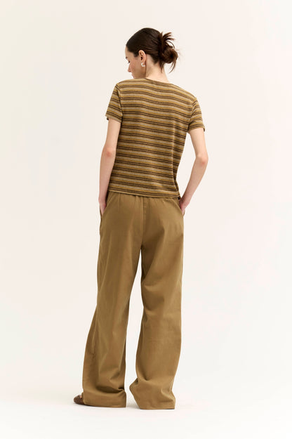 A woman shown from the back wearing a short-sleeve crewneck t-shirt with horizontal "spice variant" brown and beige stripes, paired with wide-leg brown trousers, against a plain off-white studio background.