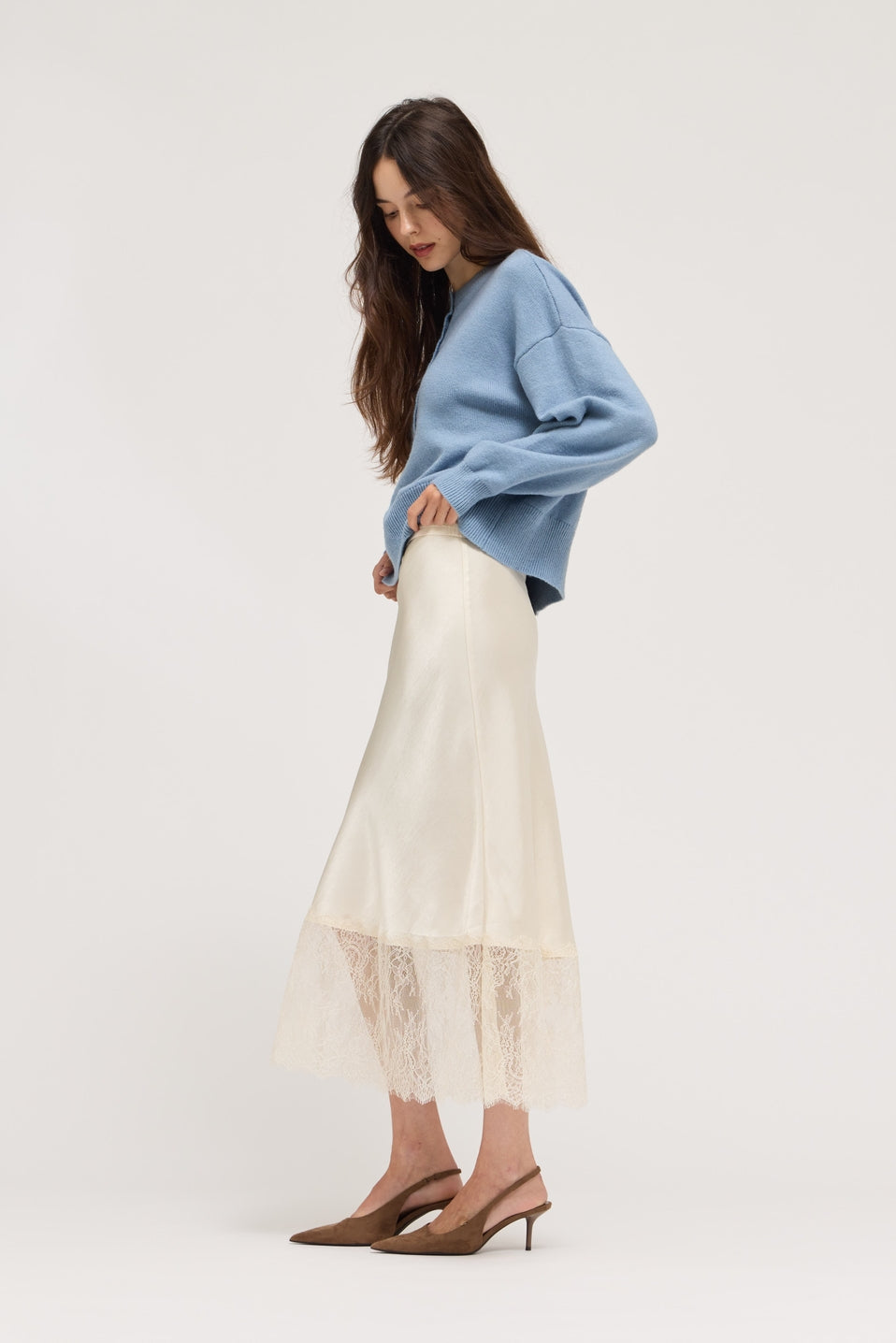 A woman modeling a light blue knit sweater and a cream satin midi skirt with a lace hem, paired with brown slingback heels, against a plain light gray studio background.