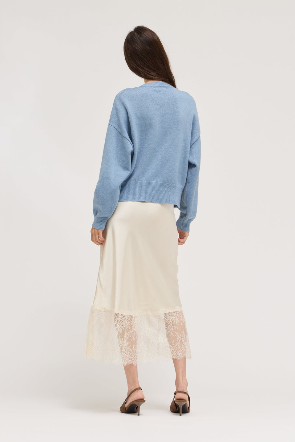 A woman is shown from behind wearing a light blue crewneck sweater and a cream-colored midi skirt with a lace hem, paired with brown heeled sandals, against a plain light gray studio background.