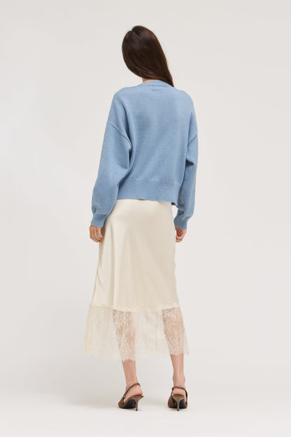 A woman is shown from behind wearing a light blue crewneck sweater and a cream-colored midi skirt with a lace hem, paired with brown heeled sandals, against a plain light gray studio background.