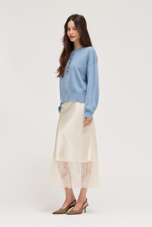 A woman modeling a light blue knit sweater and a cream satin midi skirt with a lace hem, paired with brown pointed-toe heels, against a plain light gray studio background.
