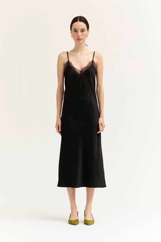 A woman modeling a black satin slip dress with a sheer black lace V-neckline and thin spaghetti straps, paired with olive green pointed-toe flats, against a plain off-white studio background.