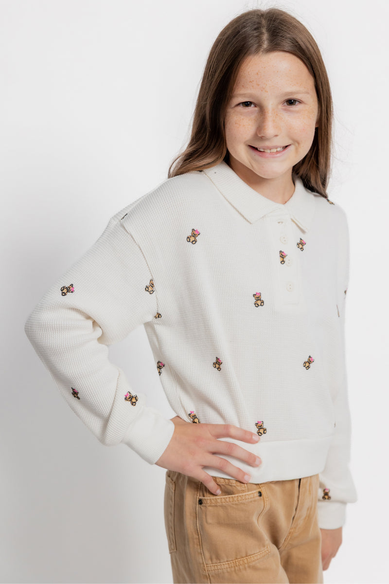 A young girl with long light brown hair smiling, wearing a cream-colored long-sleeve polo sweater with small embroidered bears and tan corduroy pants, set against a plain light gray background.