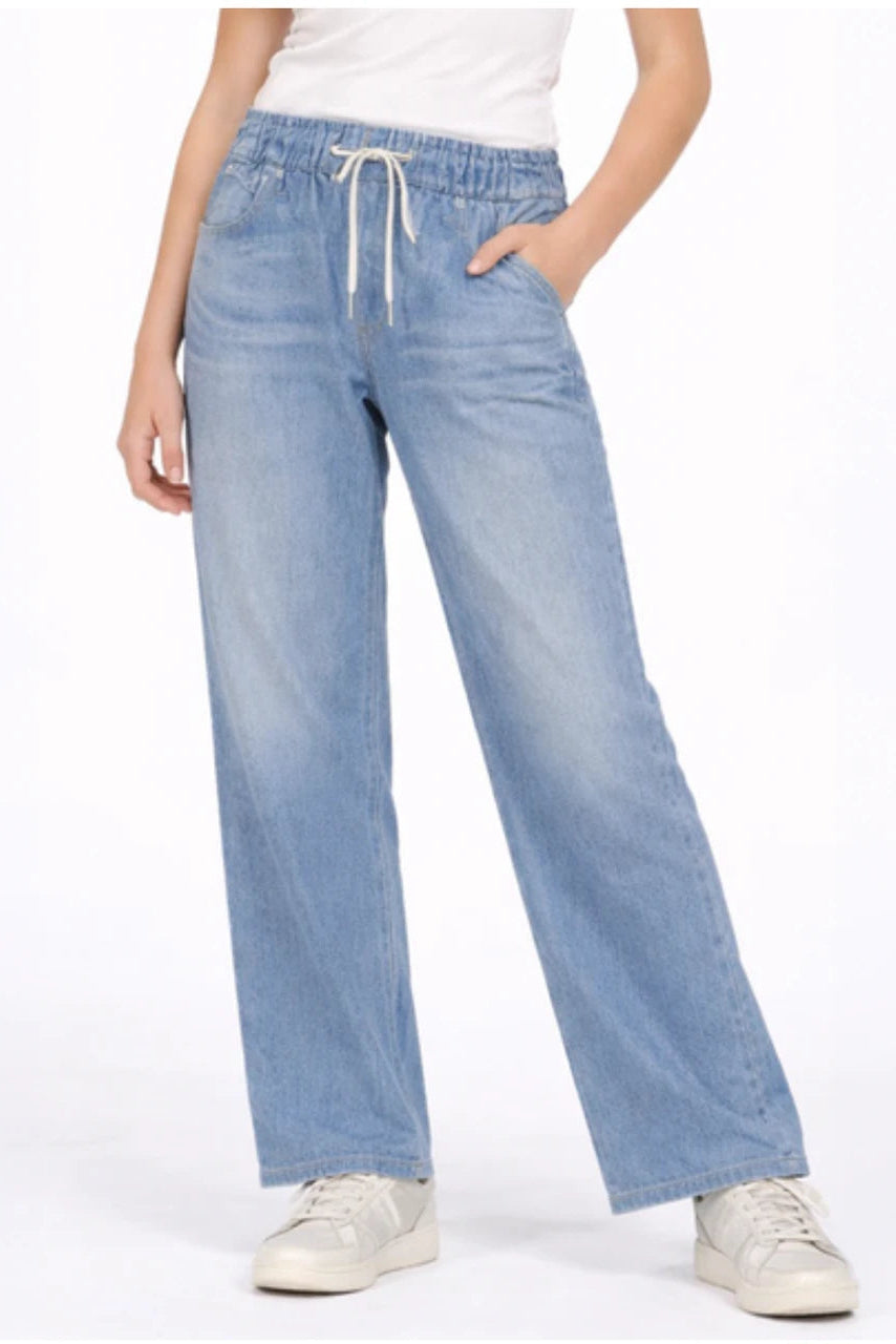 A girl wearing light blue wide-leg denim sweatpant jeans with an elastic waistband and white drawstring, paired with a white top and white sneakers, against a plain white studio background.