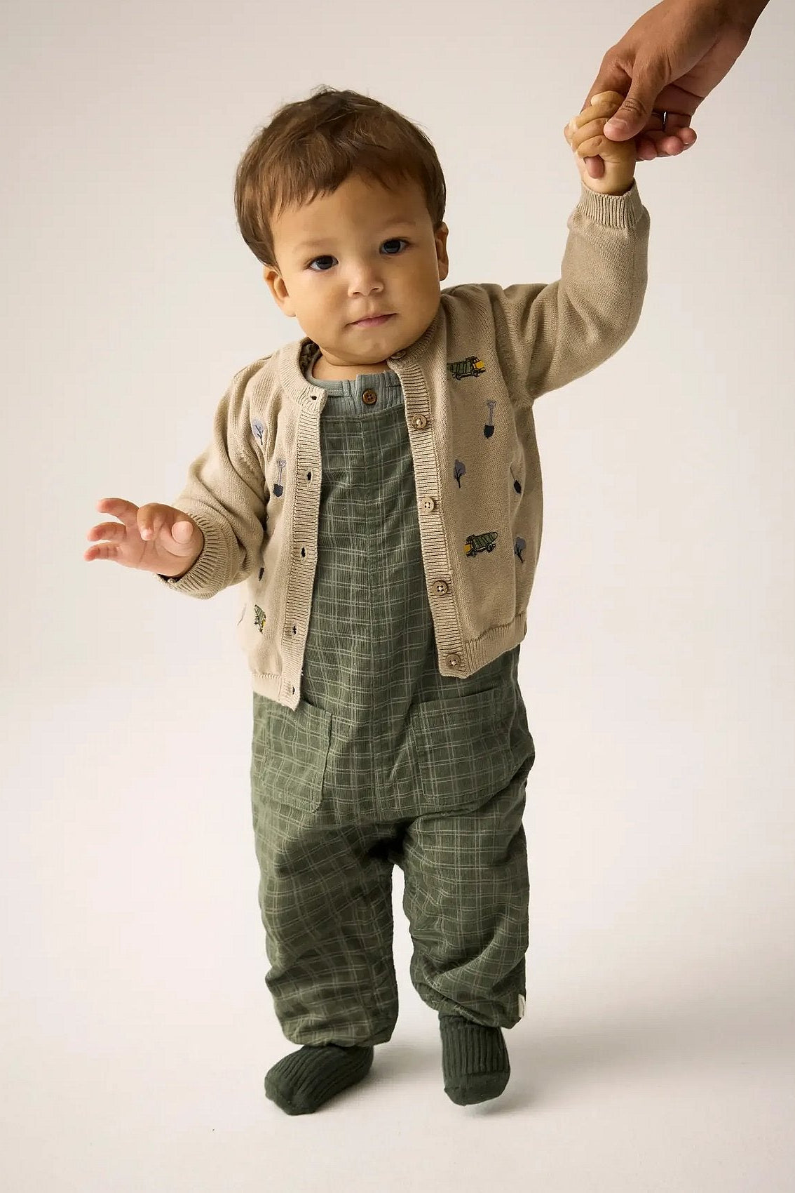 A baby wearing a beige cardigan with small embroidered motifs over  beetle green plaid overalls, standing on a white studio background while holding an adult's hand.